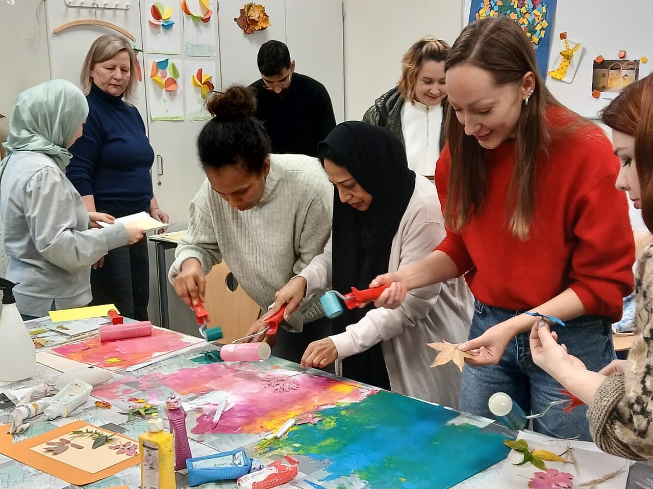 Eine Gruppe arbeitet in einem künstlerischen Prozess, es werden Pflanzenteile wie Blätter und Blüten in bunten Farben gedruckt. Im Vordergrund sieht man Blätter von verschiedenen Baumarten. Im Zentrum arbeiten mehrere Frauen sehr konzentriert an ihren Werken und im Hintergrund wird die Druckmaschine bedient.